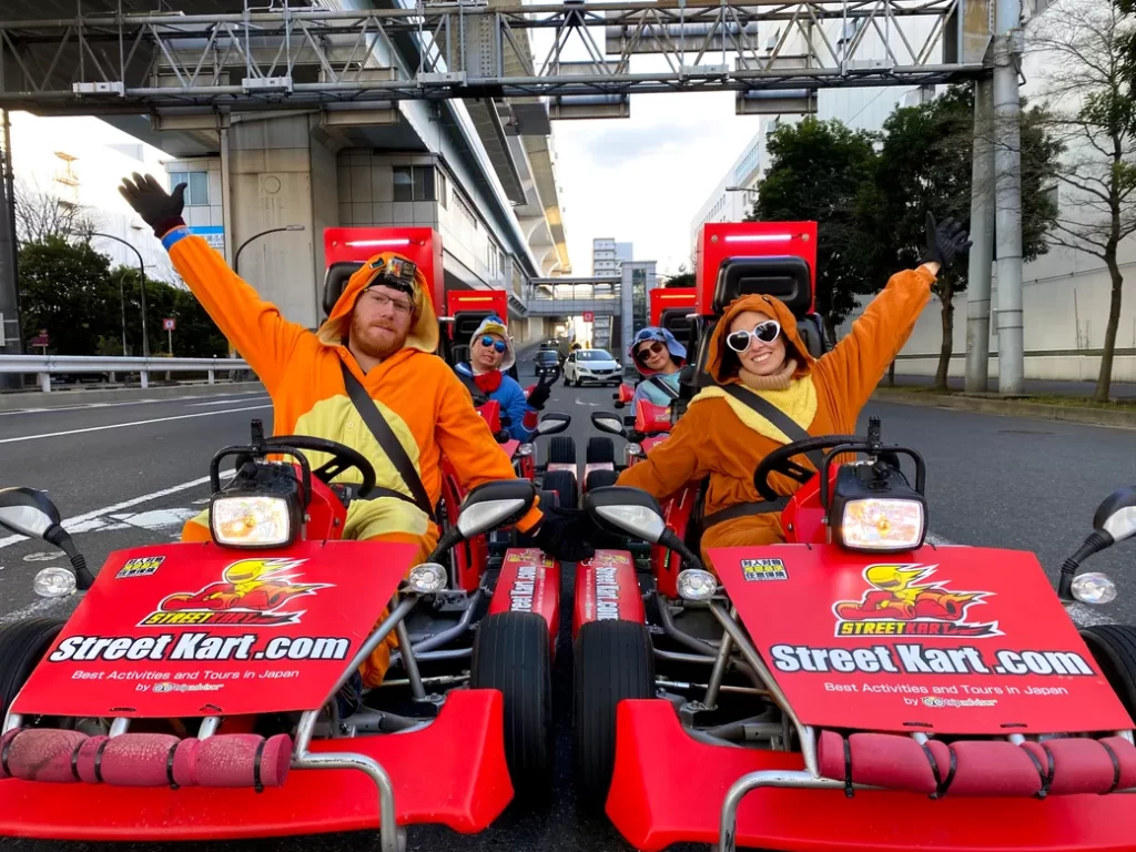 IMG_3612 - StreetKart Four people in orange animal-costume outfits ride red Street Kart go-karts on an urban street, waving at the camera.