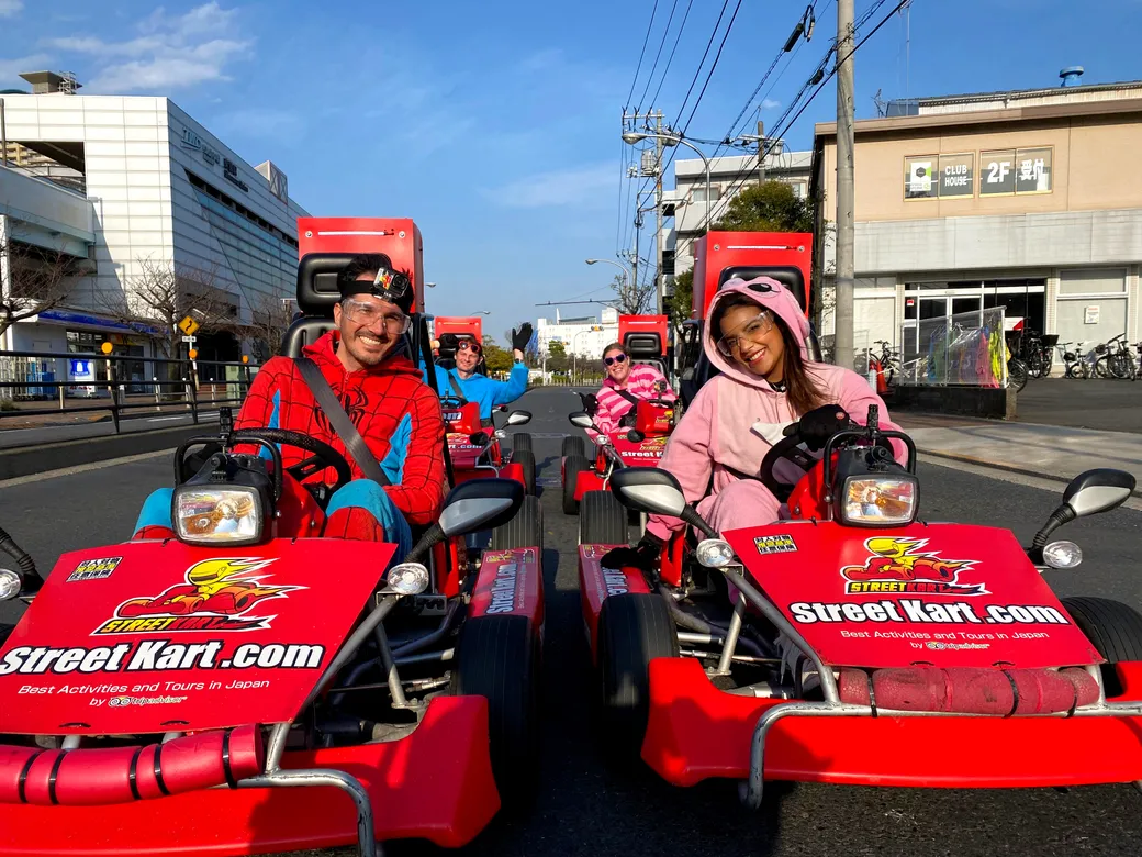 Smiling people in red go-karts with StreetKart.com branding ride on a sunny urban street, wearing colorful costumes (Spider-Man and pink onesie).